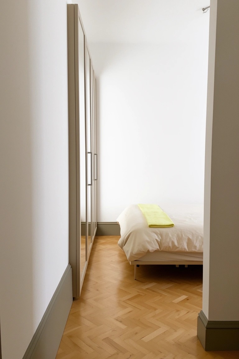 Small bedroom with bright white walls, light wood wardrobe with mirrors, parquet floor, and a low bed topped with white linens and a yellow throw