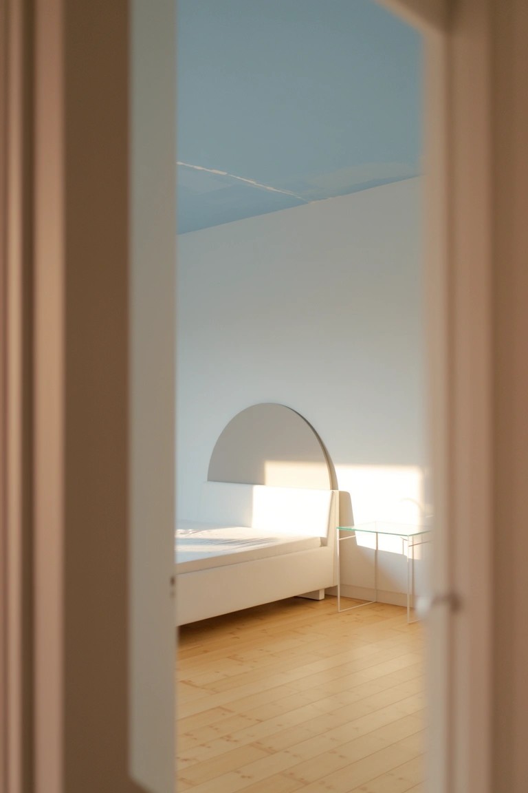 Bedroom interior viewed from doorway showing pale blue ceiling, white walls, light wood floor, and white bed with arched mirror