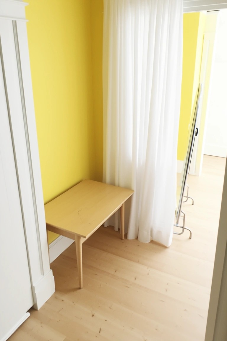 Bedroom corner featuring sunny yellow walls next to a wooden bench, white sheer curtains, and a full-length mirror on light wood floors