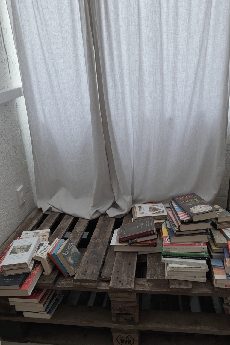 Wooden pallet shelf stacked with books against white walls and sheer curtains in a cozy bedroom nook