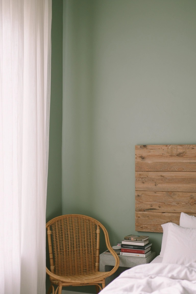 Cozy bedroom corner with pale sage green walls, wooden headboard, rattan chair, white bedding, and stacked books on the nightstand