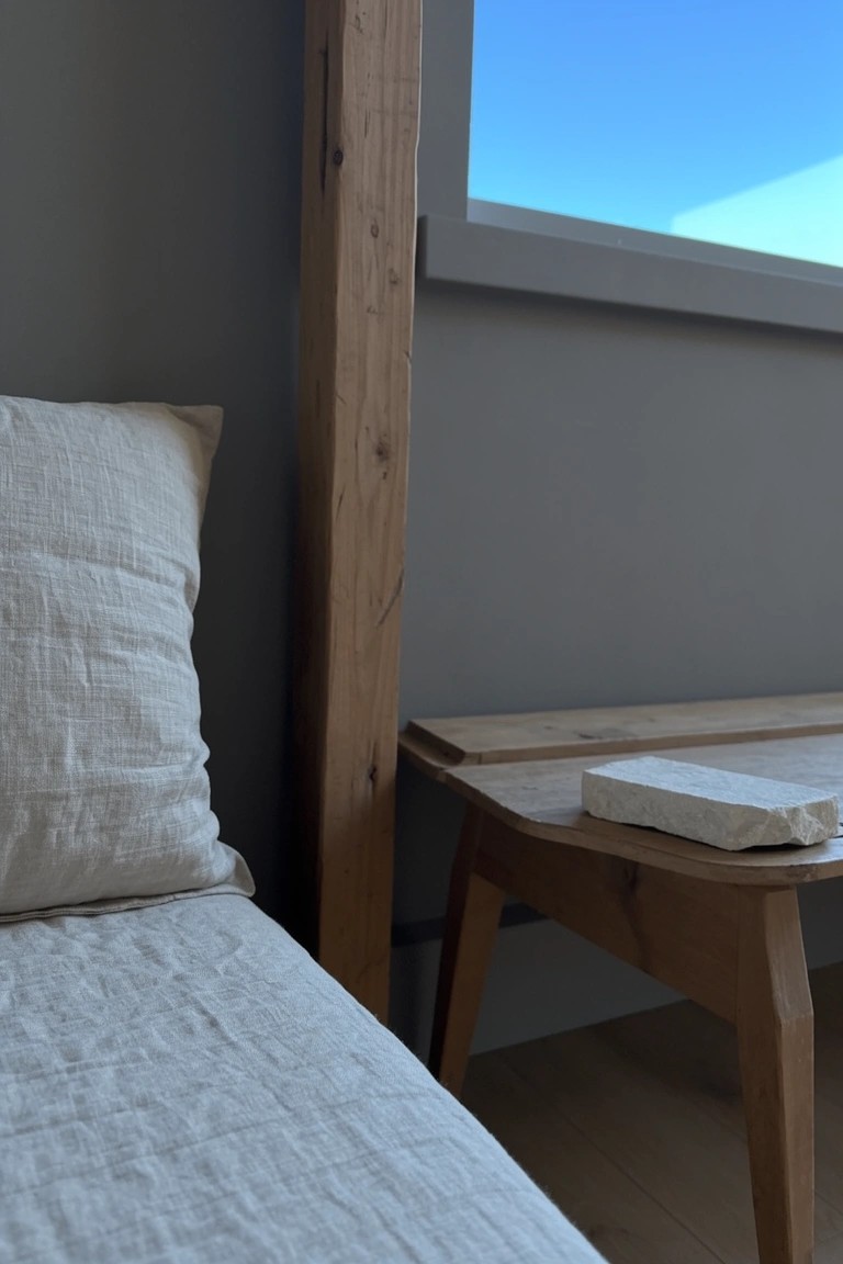 Bedroom corner featuring soft greige walls beside a wooden beam and bed with white linens, a wooden nightstand holding a book, and a window showing blue sky