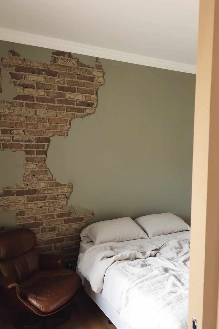 Bedroom featuring muted sage green walls beside exposed brick, a white bed with rumpled sheets, and a brown leather armchair