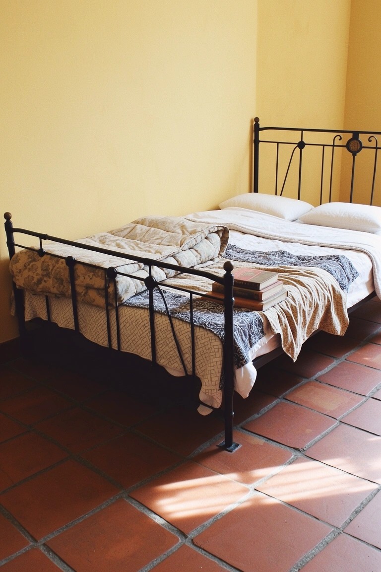 Bedroom corner with pale butter yellow walls, black wrought-iron bed frame piled with books and rumpled neutral bedding, terracotta tile floor, soft sunlight streaming in