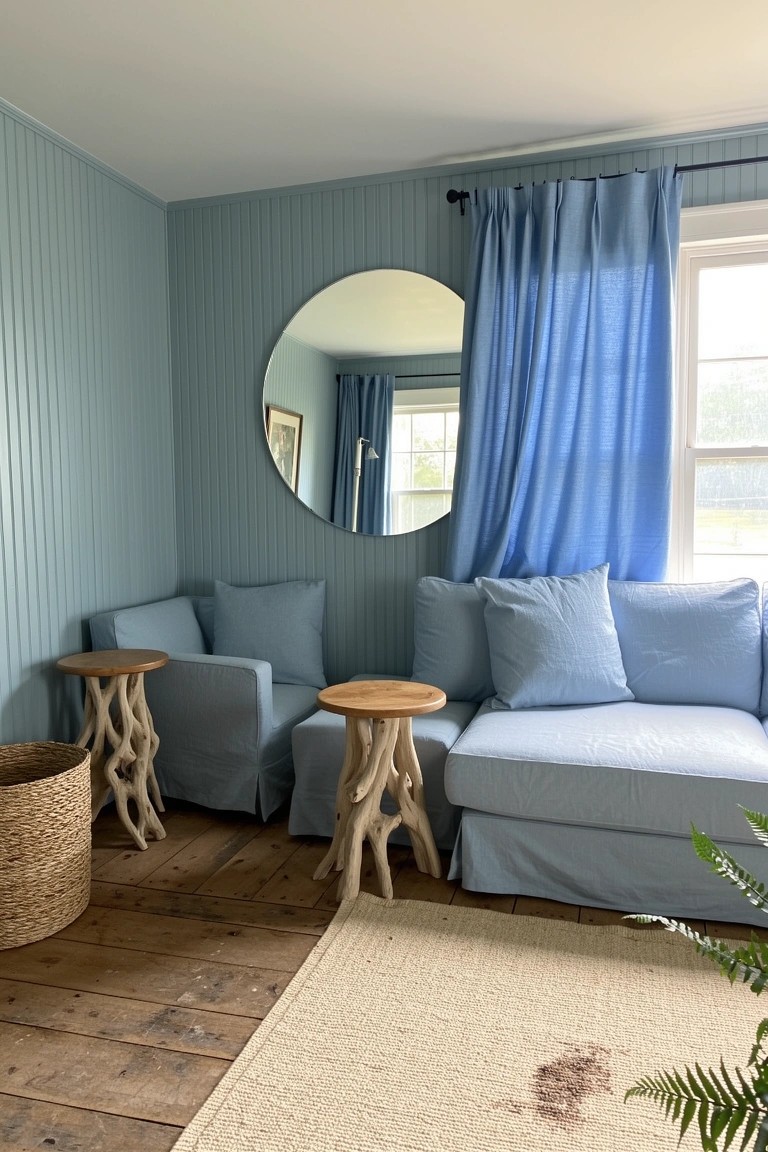 Light blue slipcovered sofa and chairs with wood stump side tables in a pale blue planked wall corner, round mirror and blue curtains on window