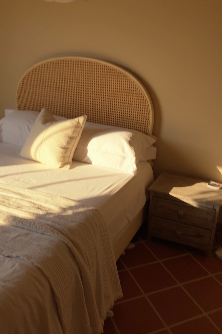 Sunlit bedroom featuring warm beige walls, curved rattan headboard, white pillows and duvet, wooden nightstand, and red terracotta floor tiles