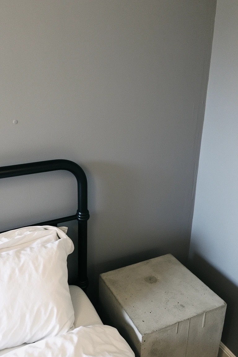 Pale gray walls in a simple bedroom with black metal bed frame, white bedding, and concrete side table