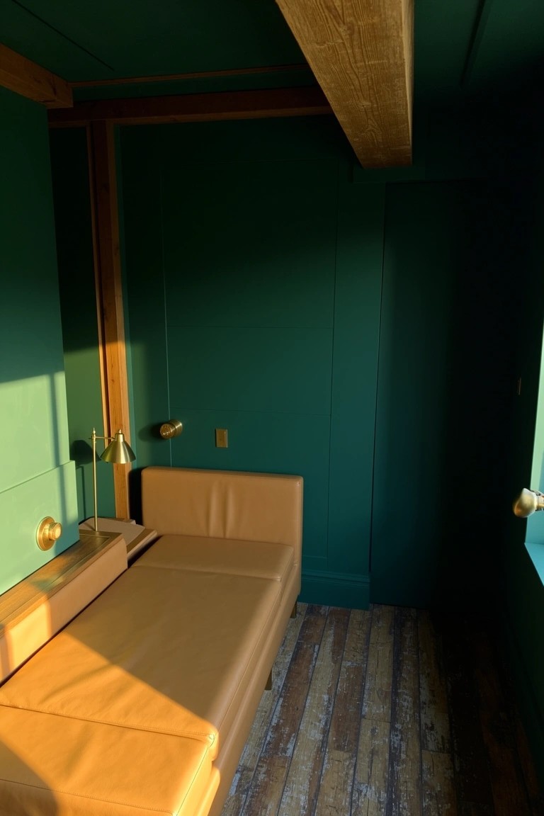 Small bedroom nook with deep green walls and ceiling, tan leather daybed, wooden beams and flooring, brass lamps and warm sunlight