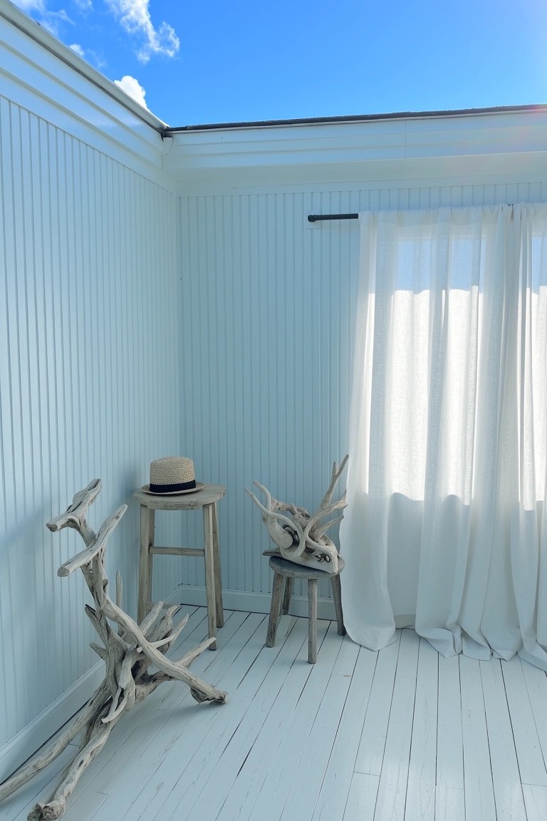 Pale blue shiplap walls in a bright bedroom corner with driftwood stools, a straw hat, white wood floors, and sheer curtains