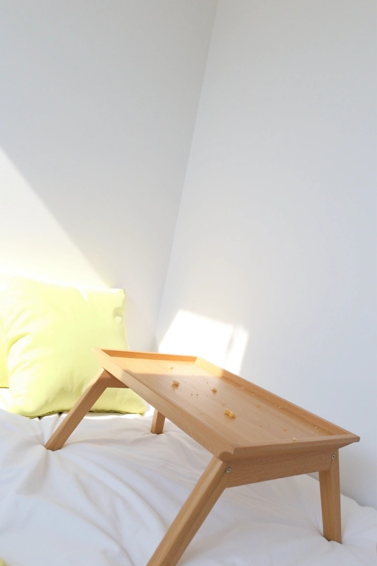 Minimalist bedroom corner with crisp white walls, wooden lap desk on white bedding, and lime green pillow.