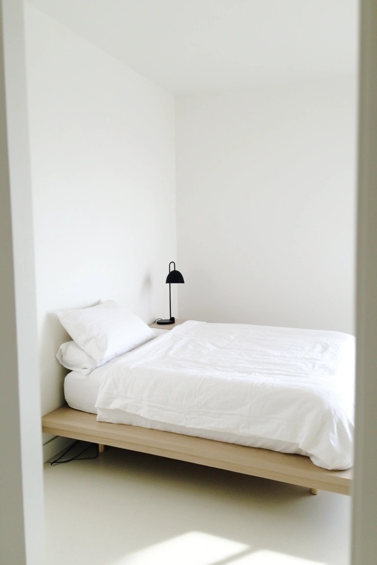 Minimalist bedroom with crisp white walls, light wood platform bed, white bedding, and black lamp on nightstand