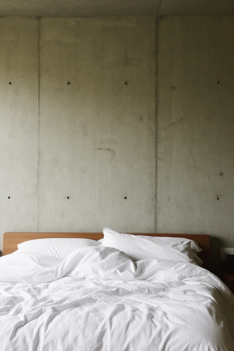 Minimalist bedroom featuring exposed pale gray concrete walls, a simple wooden bed frame, and rumpled white bedding for a clean modern vibe.