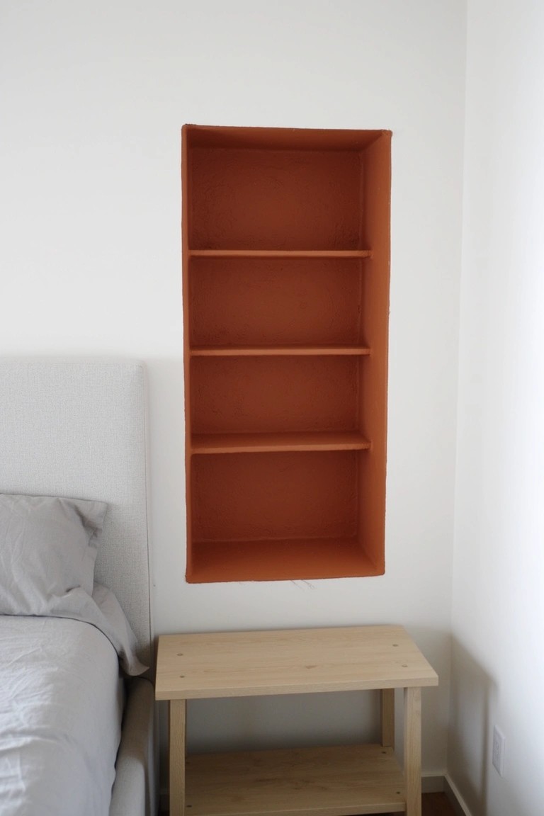 Recessed shelving unit painted warm terracotta orange, built into a white wall in a minimalist bedroom with gray bedding and wooden nightstand