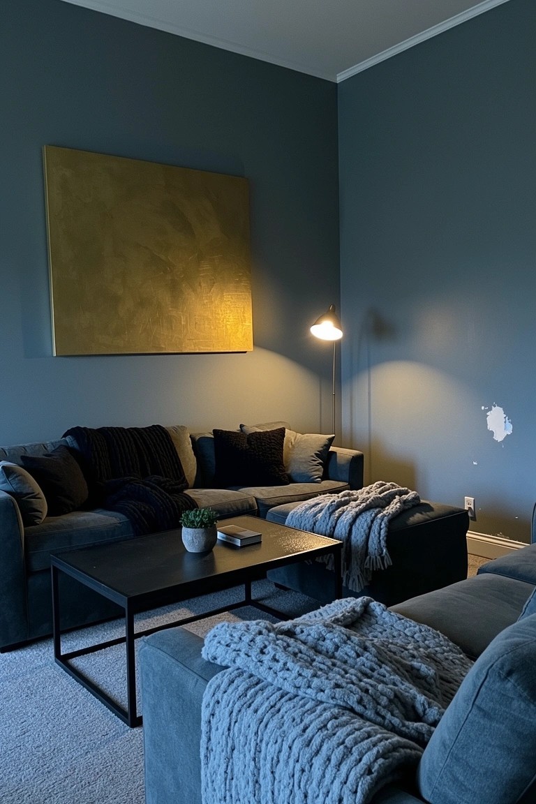 Cozy living room corner featuring deep navy walls, gray L-shaped sofa with dark cushions and chunky knit throw, black metal coffee table with plant, large gold abstract painting, and warm arched floor lamp.