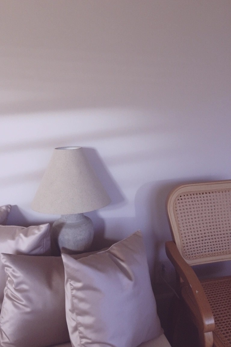 Bedroom corner with pale greige walls, stone-based lamp, satin pillows on bed, and rattan chair in soft light