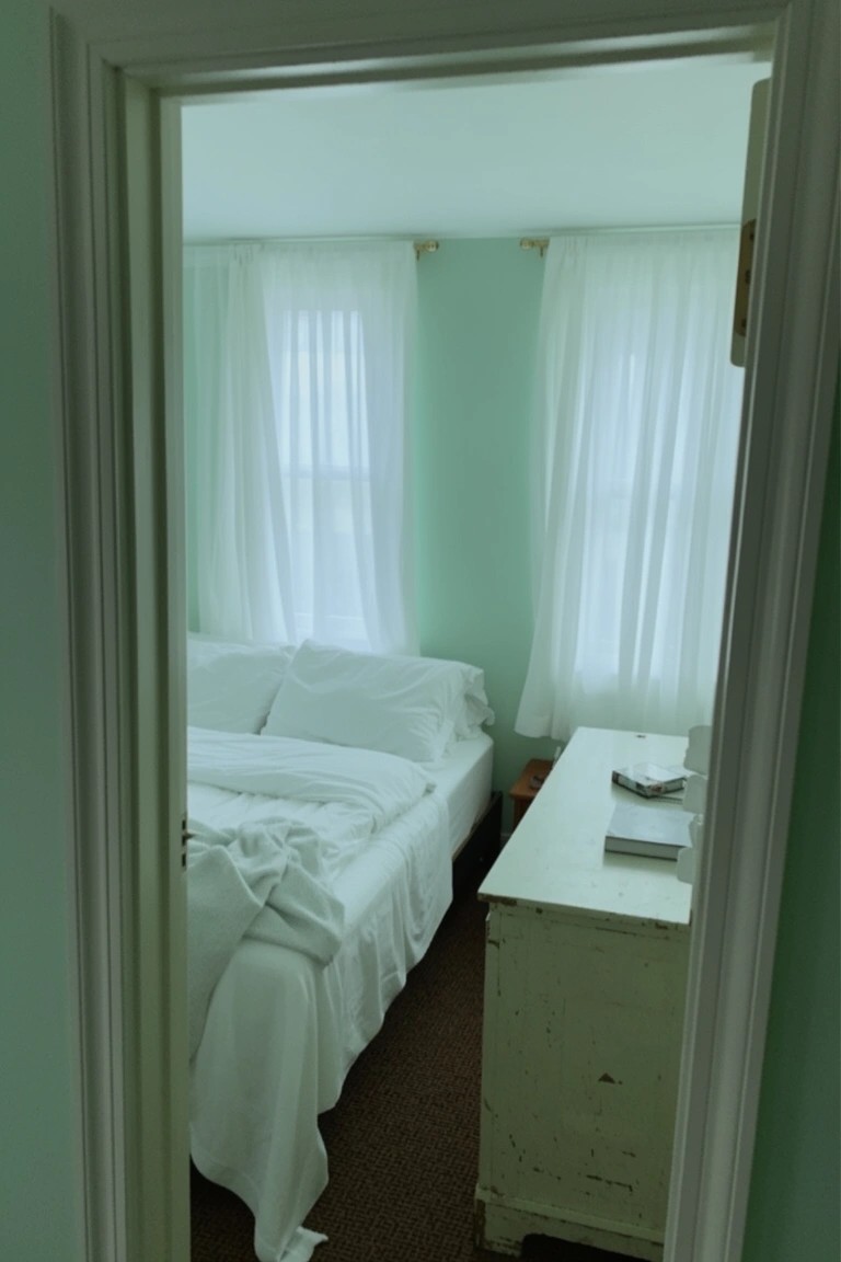 Bedroom interior with pale mint green walls, white curtains and bedding, wood dresser by the bed