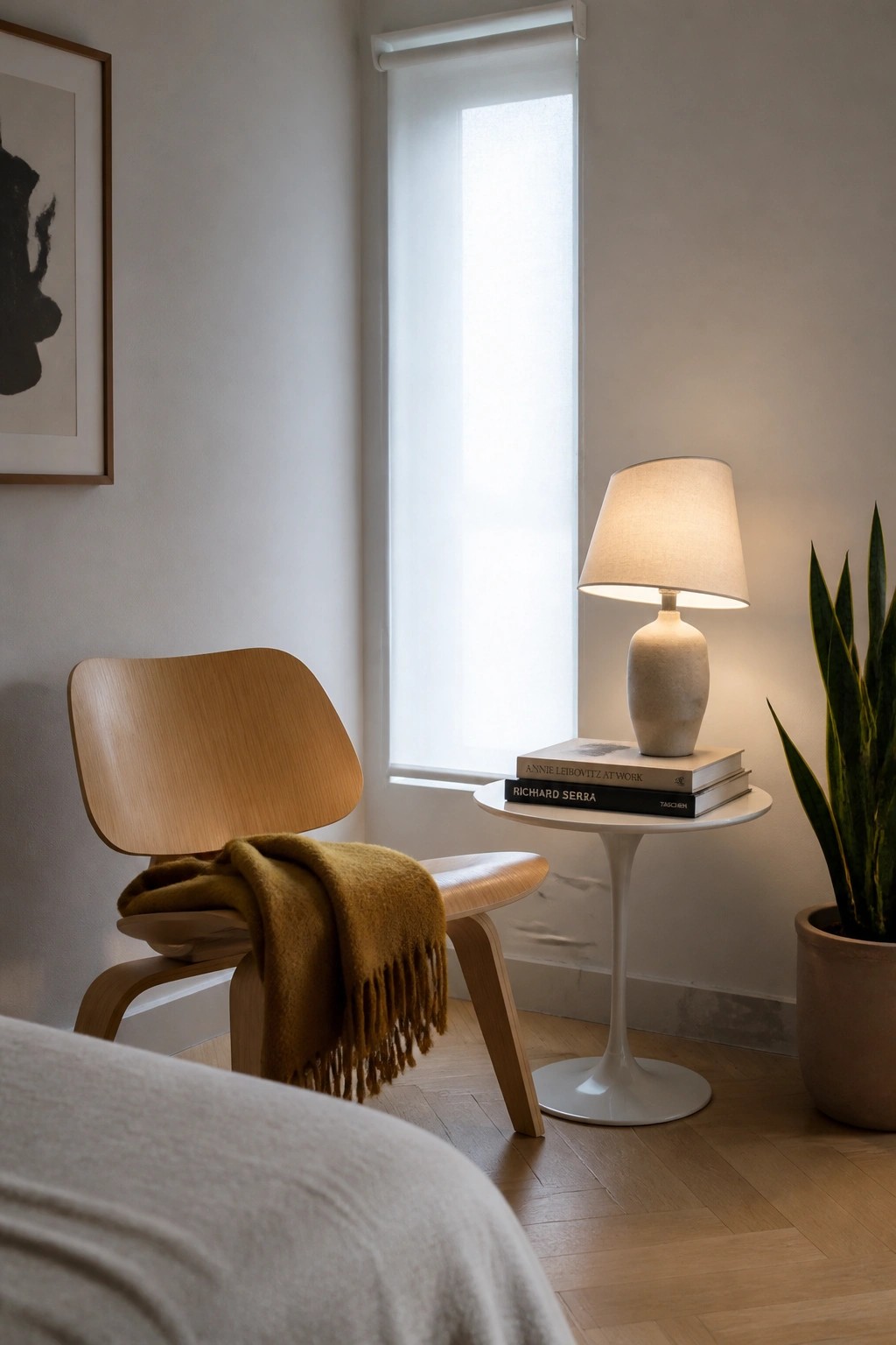 A light wood lounge chair with a mustard throw blanket sits beside a round white side table holding a lamp and books in front of a window.