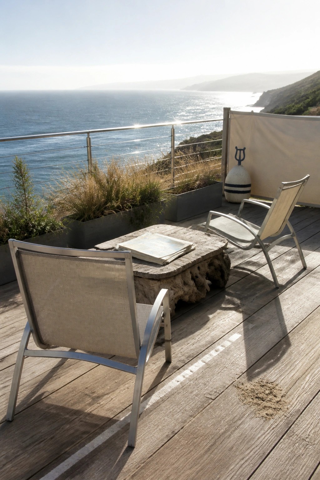 Two metal frame lounge chairs with mesh seats sit on a wooden deck beside a rustic table, overlooking the ocean.
